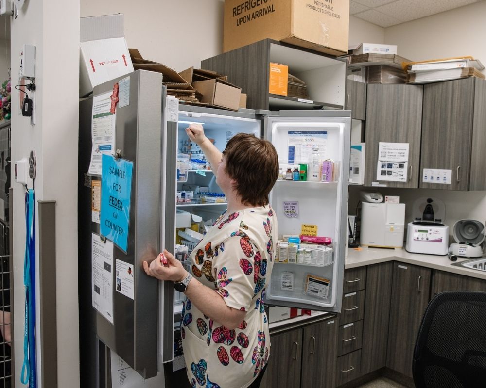A person in a patterned scrub top reaches into an open refrigerator filled with medication bottles and vials.