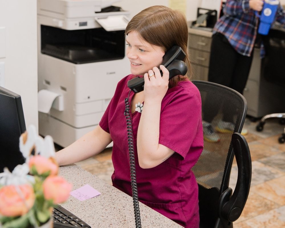 A person in a purple scrub top talks on a black phone while sitting at a reception desk.