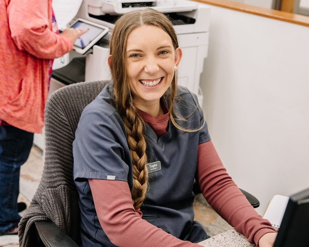 Image 2 A woman in scrubs smiles while sitting at a reception desk with a computer