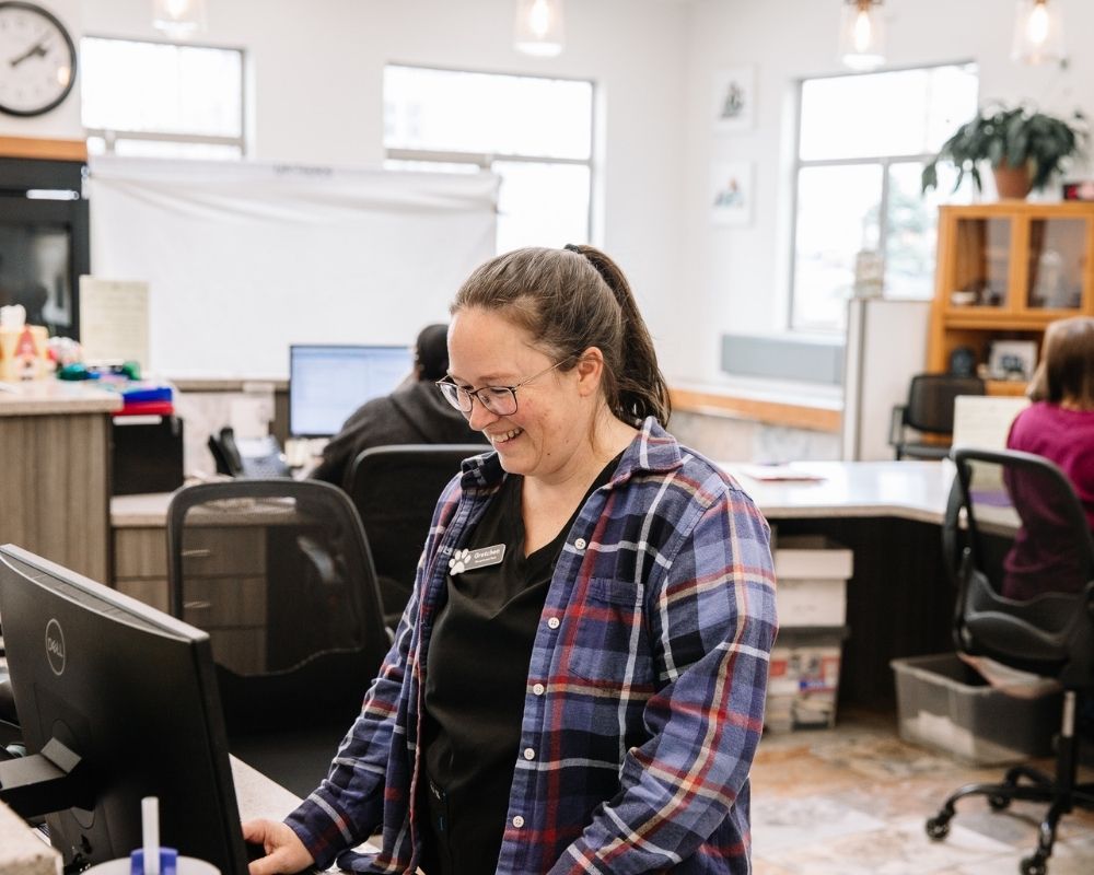 Image 2 A person wearing glasses and a plaid shirt smiles while looking at a computer monitor at a reception desk.