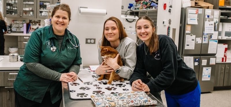 A veterinarian and two women with a small pet in a veterinary clinic examination room.